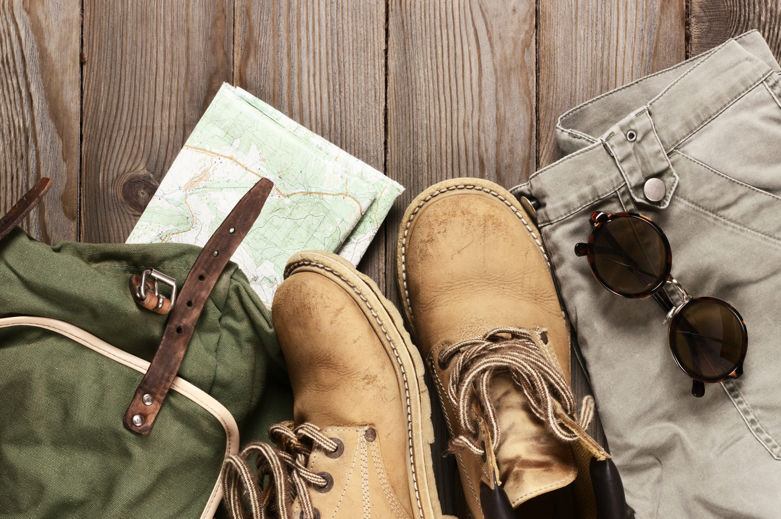 Travel accessories set on wooden background: old hiking leather boots, pants, backpack, map and sunglasses. Top view point.